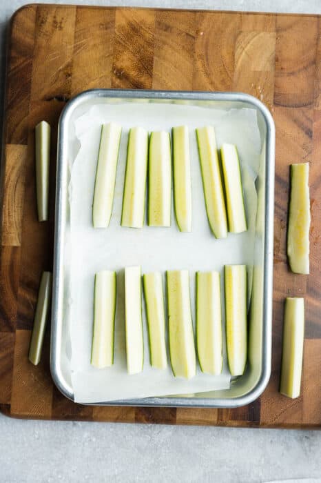 Top view of uncooked zucchini pieces on a baking sheet on a wooden cutting board