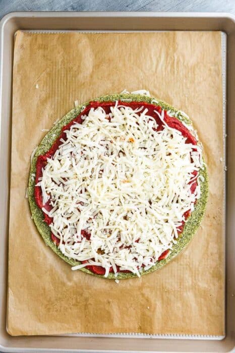 Top view of unbaked zucchini Pizza Crust with tomato sauce and cheese on brown parchment paper on a baking sheet