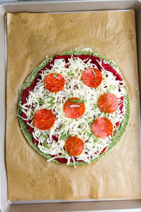 Top view of unbaked zucchini Pizza Crust with tomato sauce, cheese and pepperoni on brown parchment paper on a baking sheet