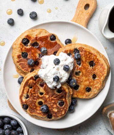 A plate of three fluffy blueberry pancakes topped with fresh blueberries with whipped cream on a white plate